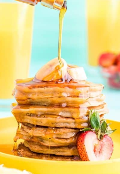 Close up photo of a stack of banana pancakes on a yellow plate with maple syrup being poured over the top.