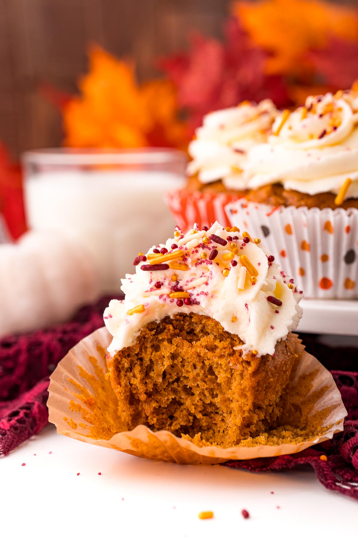 A pumpkin cupcake with a bite taken out of it on a white table with a cake stand with more cupcakes and a glass of milk in the background.