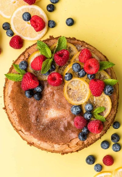 Overhead photo of lemon olive oil cake topped with berries and candied lemons on a yellow surface.