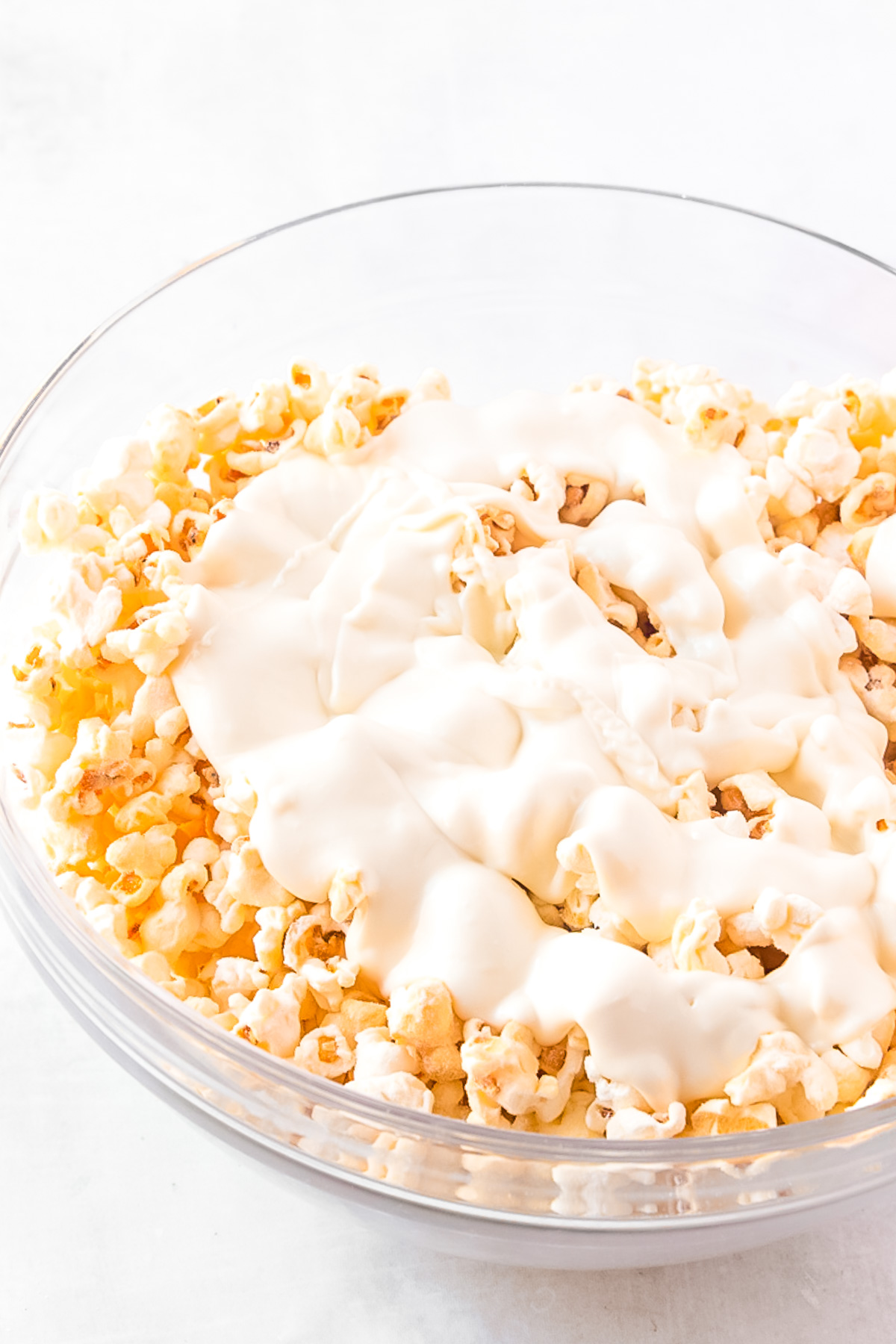 Melted white chocolate on top of popcorn in a mixing bowl.