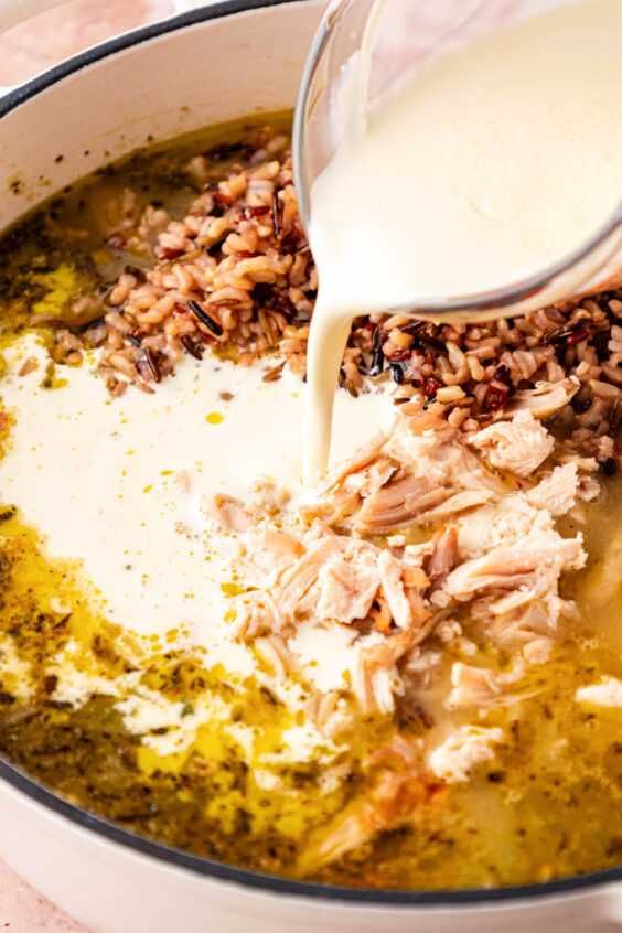 Cream, shredded chicken, and cooked wild rice being added to a pot of soup.