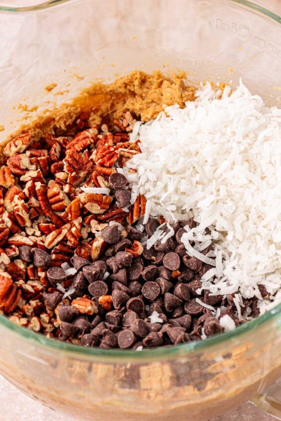 Coconut, chocolate chips, and pecans being added to a mixing bowl with cookie dough.