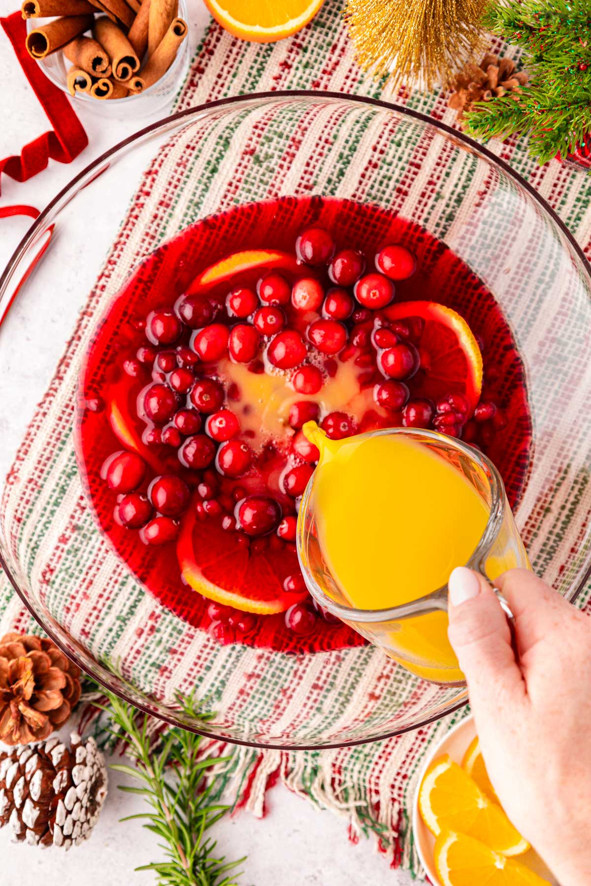 A woman's hand pouring orange juice from a measuring cup into a punch bowl with other ingredients in it.