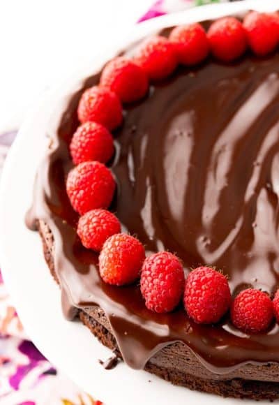 Close up photo of a chocolate cake topped with ganache and raspberries on a white cake stand.