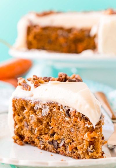 Slice of carrot cake on a white plate with a fork. Full cake on a cake stand in the background.