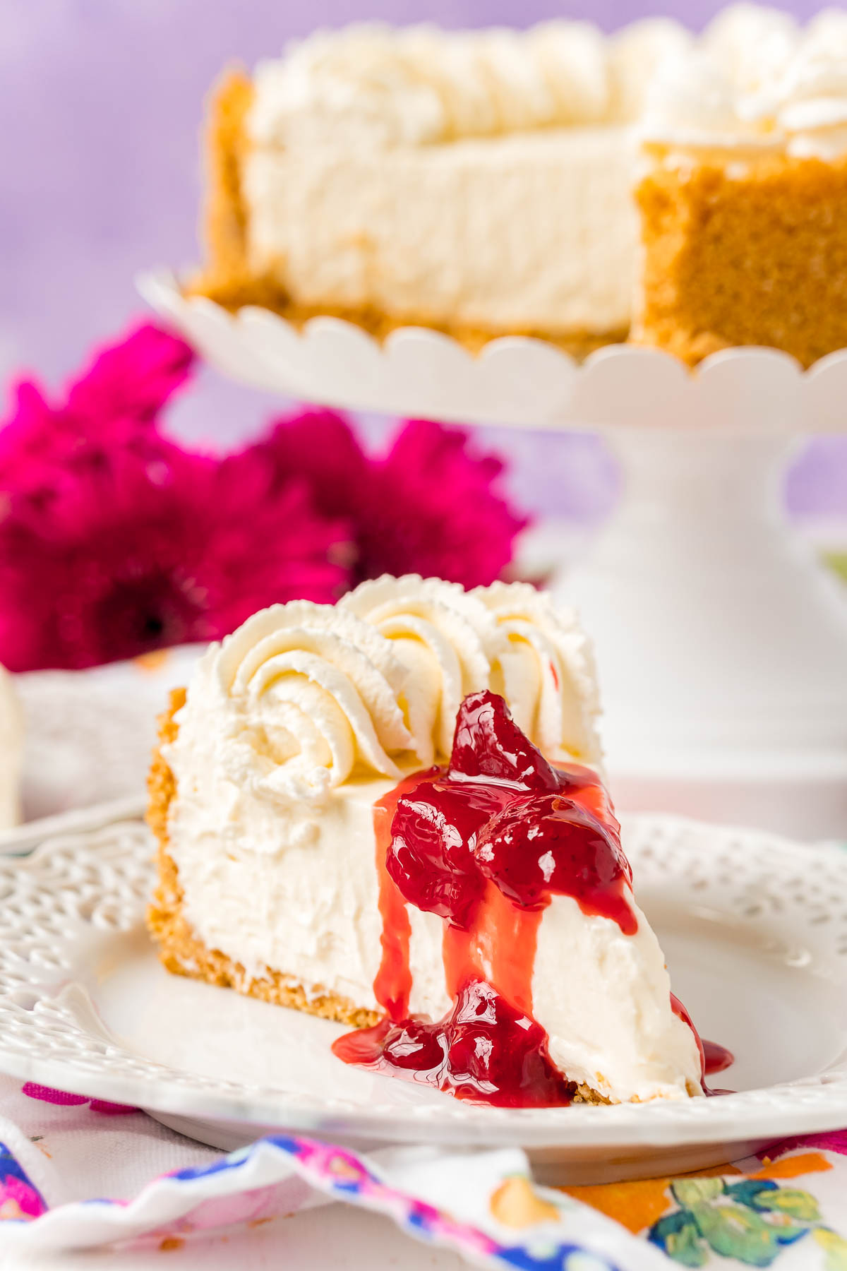 Cheesecake slice on white plate topped with whipped cream and strawberry sauce.