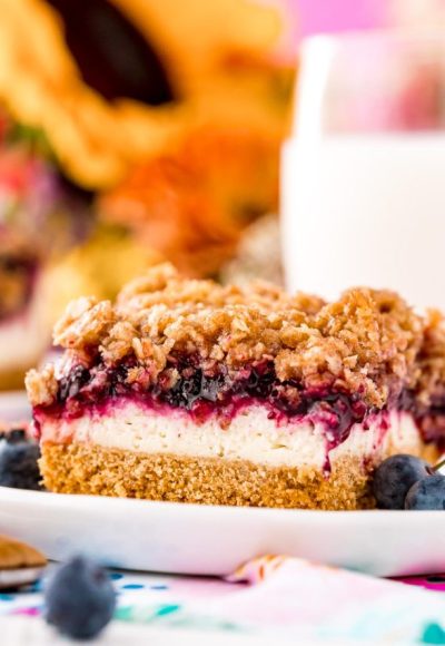 Close up photo of a slice of blueberry cheesecake bars on a white plate with milk and sunflowers in the background.