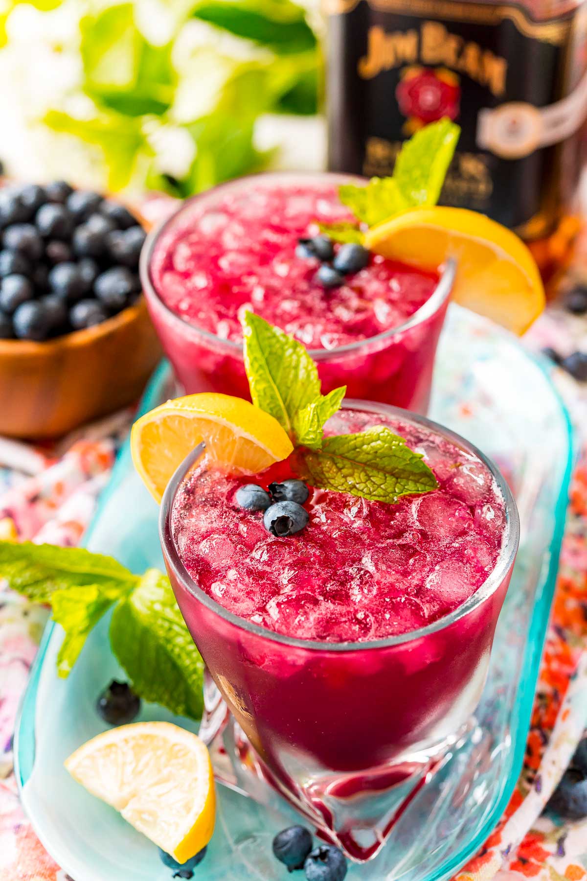 Blueberry bourbon smash on a blue serving tray with lemons, mint, and blueberries scattered around.