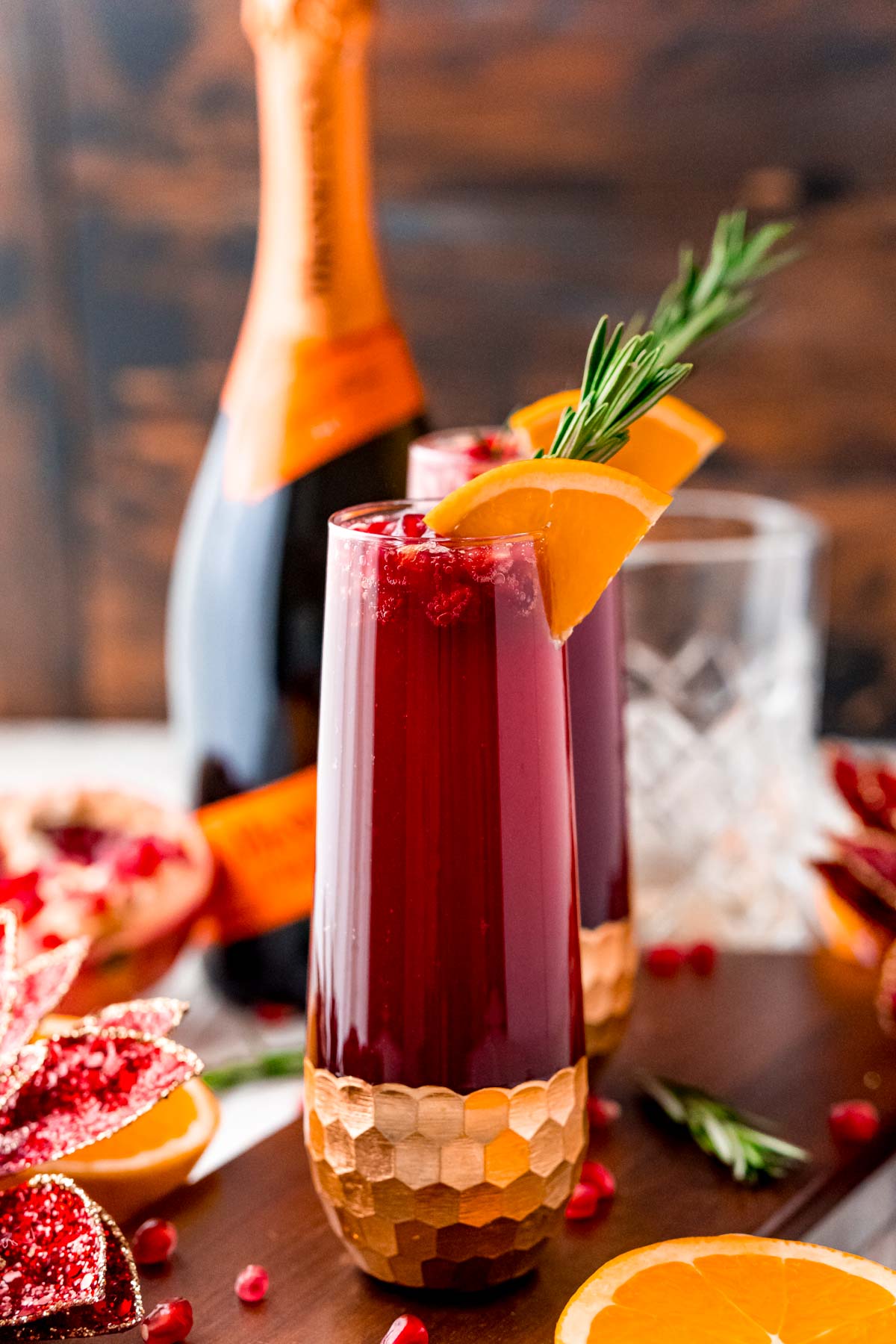 Close up photo of a red mimosa garnished with orange slice and rosemary. Prosecco bottle and pomegranates in the background.