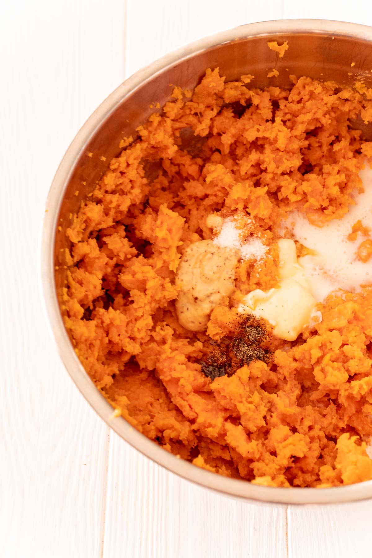 Mashed sweet potatoes in a metal mixing bowl with buttermilk, mustard, salt, and pepper.