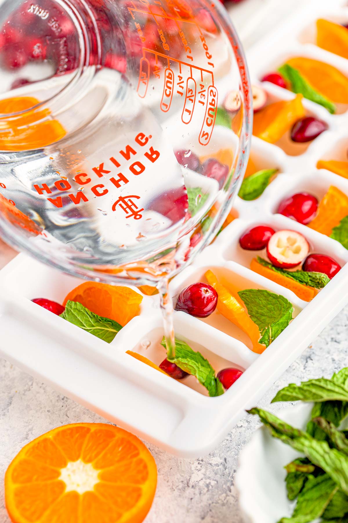 Water being poured into ice cube trays with cranberries, mint, and orange.