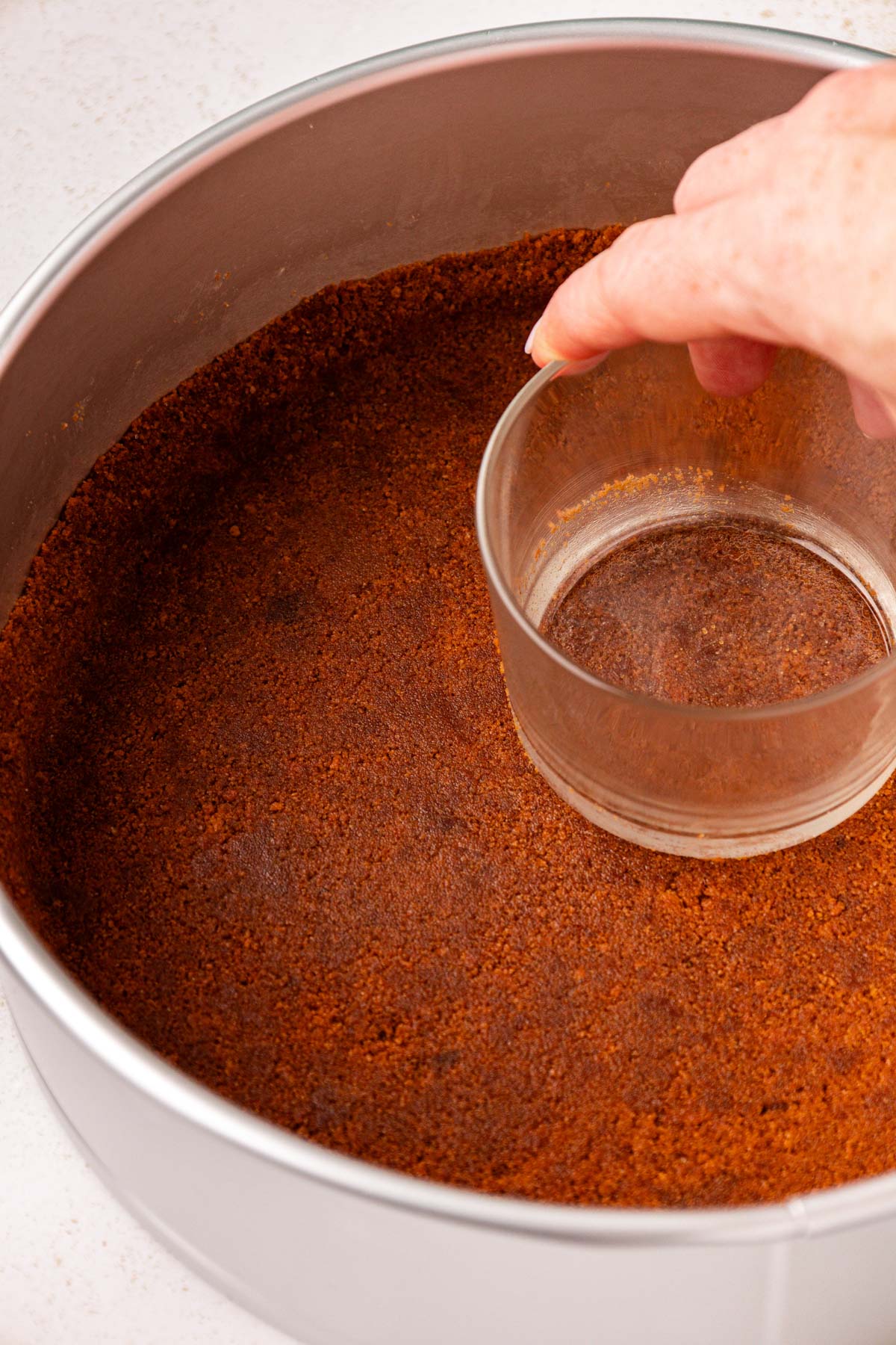 A woman's hand pressing a glass into the bottom of a springform pan to make a cookie crust.