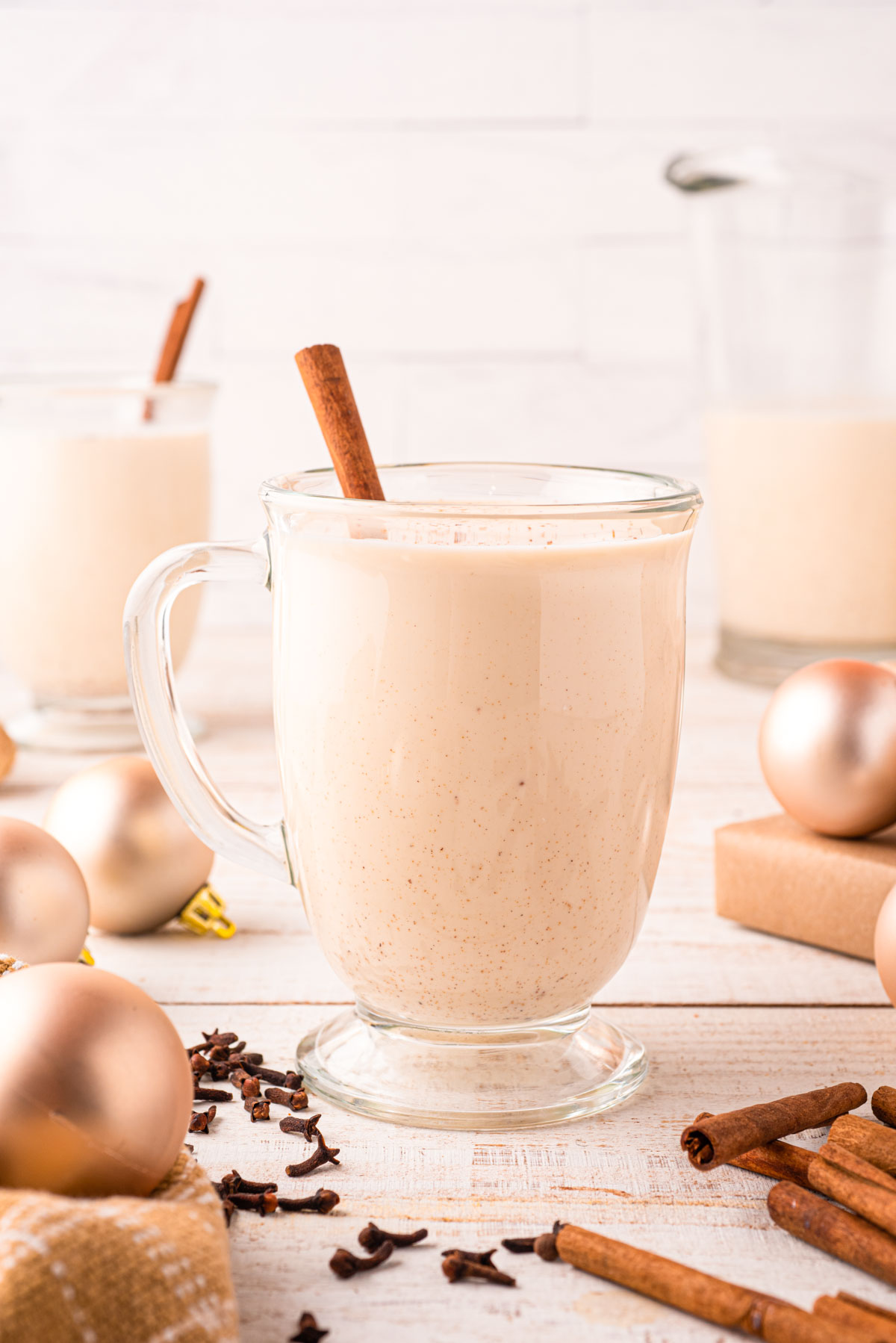 A clear glass mug of eggnog on a table with holiday decorations and spices.