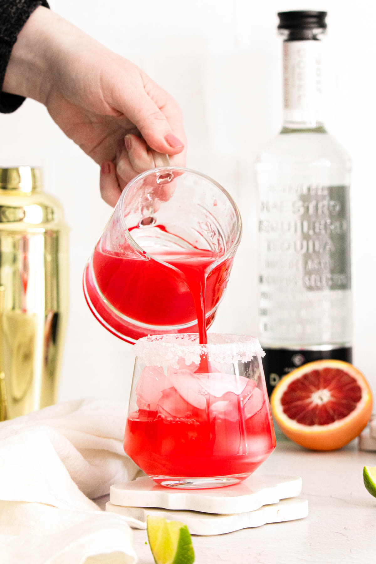 A pitcher pouring a blood orange margarita into a cocktail glass with ice.