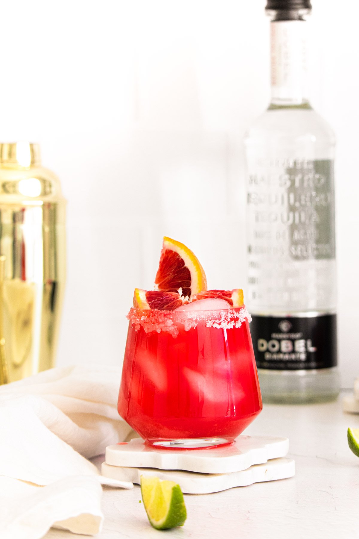 A blood orange margarita with a bottle of vodka and a cocktail shaker in the background.