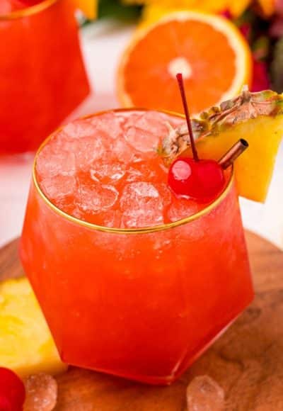 Close up photo of a gold rimmed glass with a bahama mama in it garnished with a maraschino cherry and a pineapple wedge.