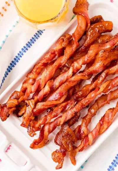 Close up photo of twisted bacon on a white plate with a glass of orange juice next to it.