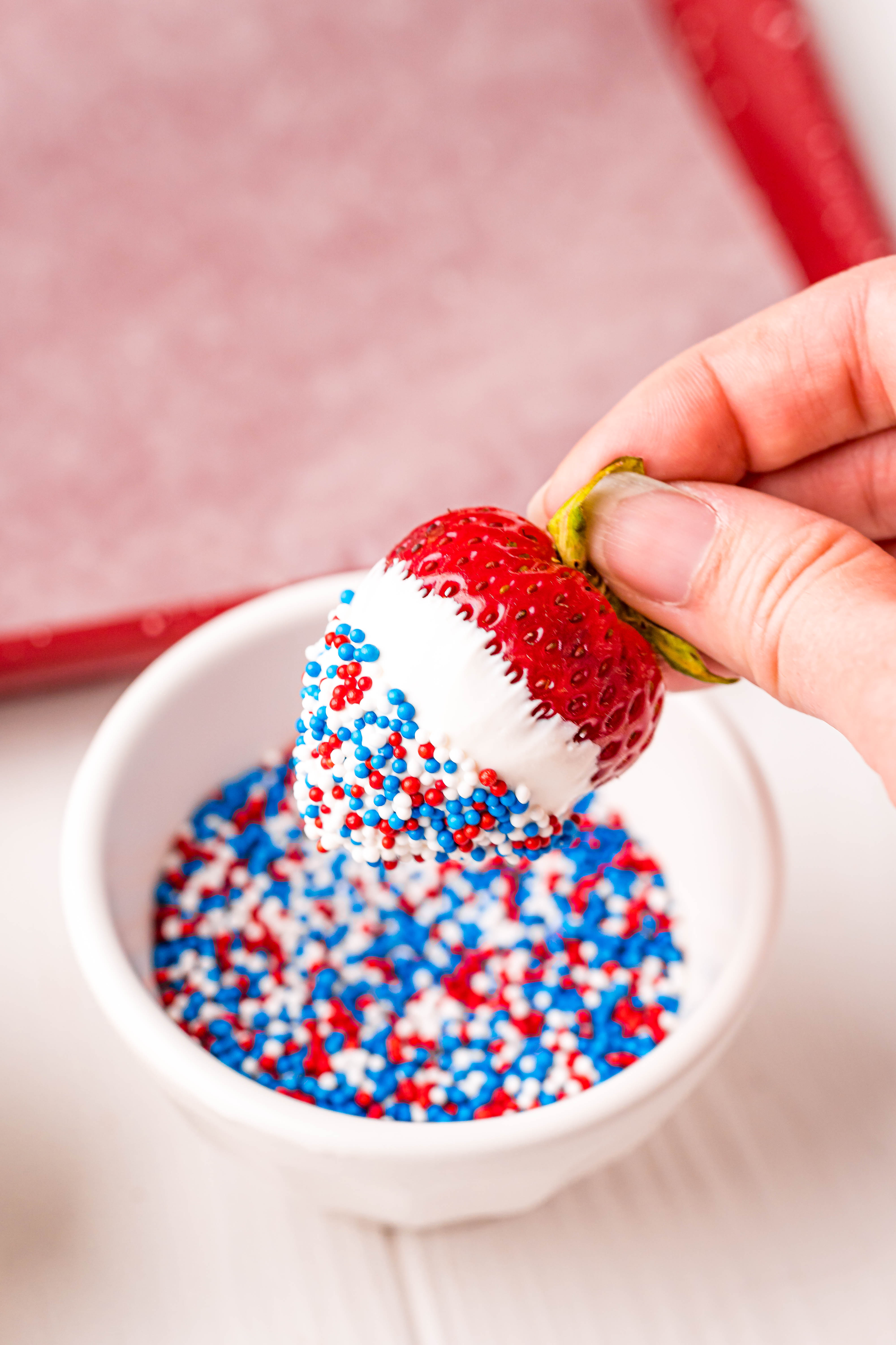 A woman's hand dipping a white chocolate covered strawberry in red, white, and blue sprinkles.