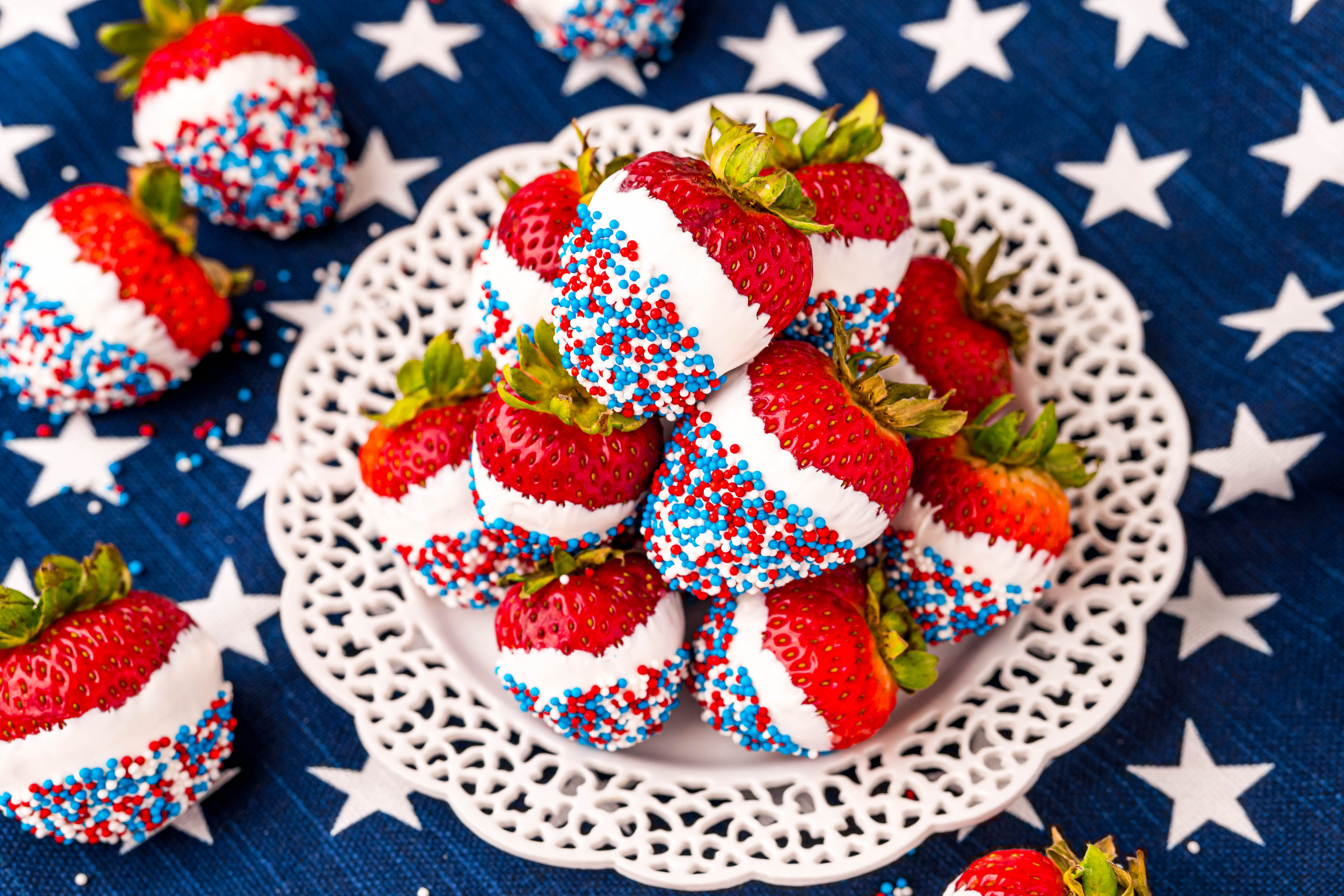 Red, white, and blue dipped strawberries on a white plate on a blue and white star printed napkin.