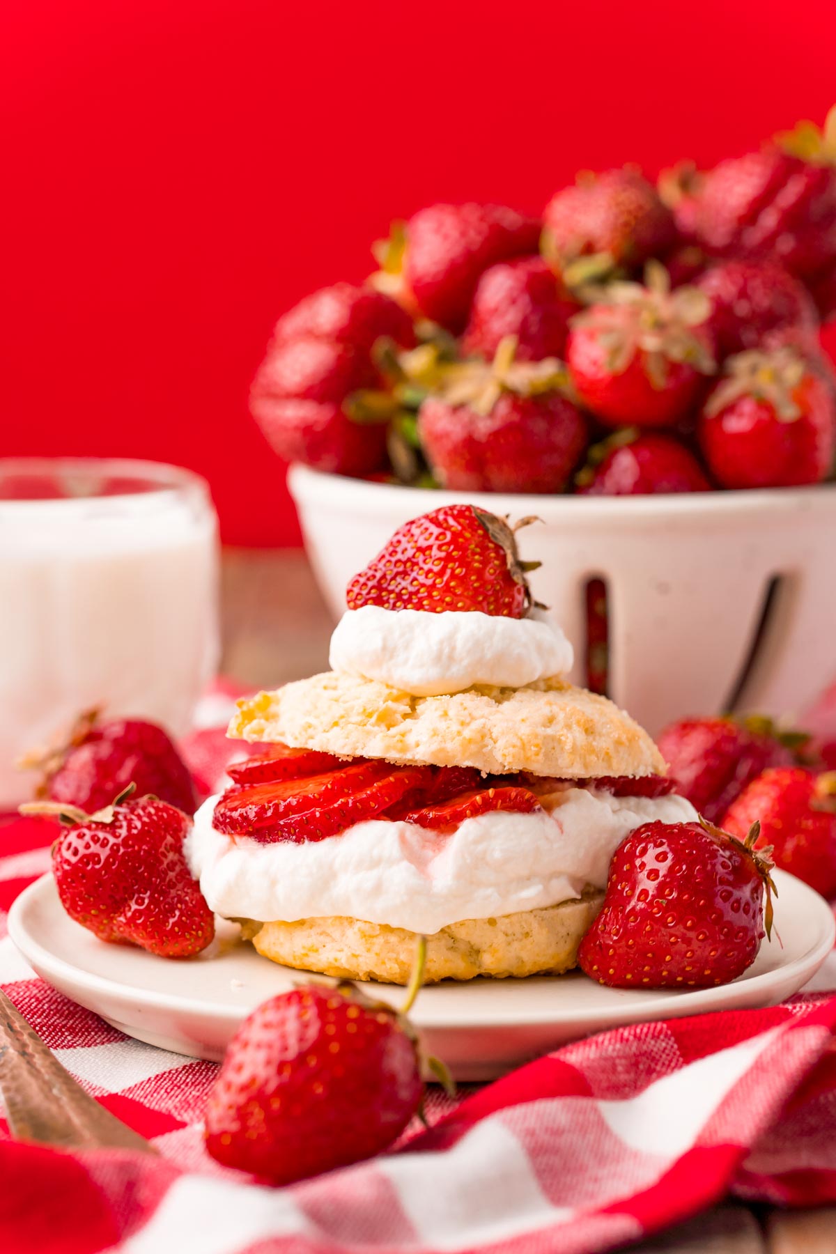 Straight on photo of strawberry shortcake on a white plate on a red and white checkered napkin.