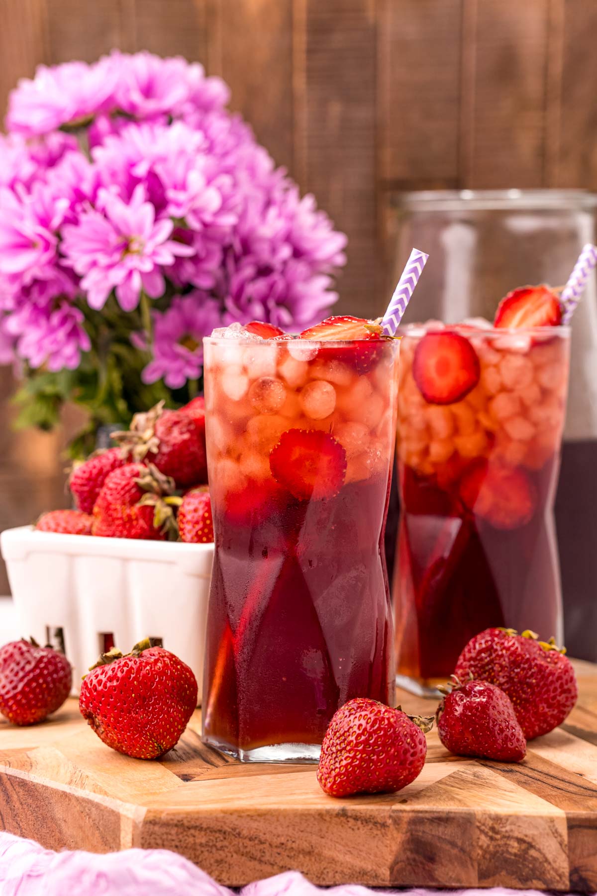 Photo of two glasses on strawberry sweet tea on a wooden board with strawberries scattered around and purple flowers in the background.