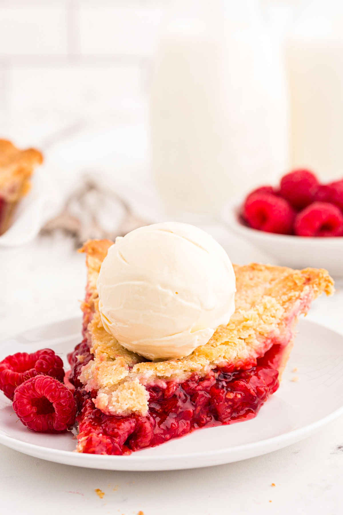 A slice of raspberry pie on a white plate with a scoop of vanilla ice cream on top.