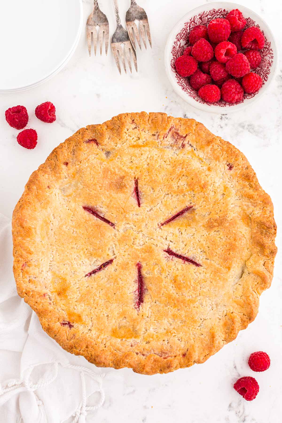 Overhead photo of a whole raspberry pie on a white table with raspberries around it.