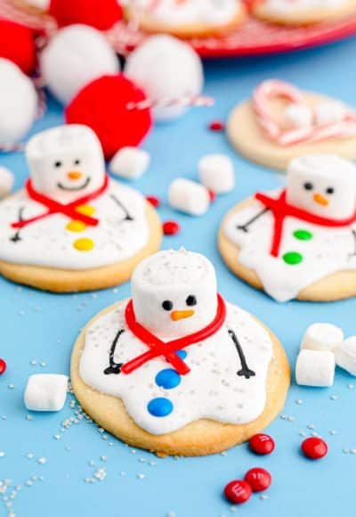 Melted Snowman Cookies on a blue surface with candy and marshmallows scattered around.