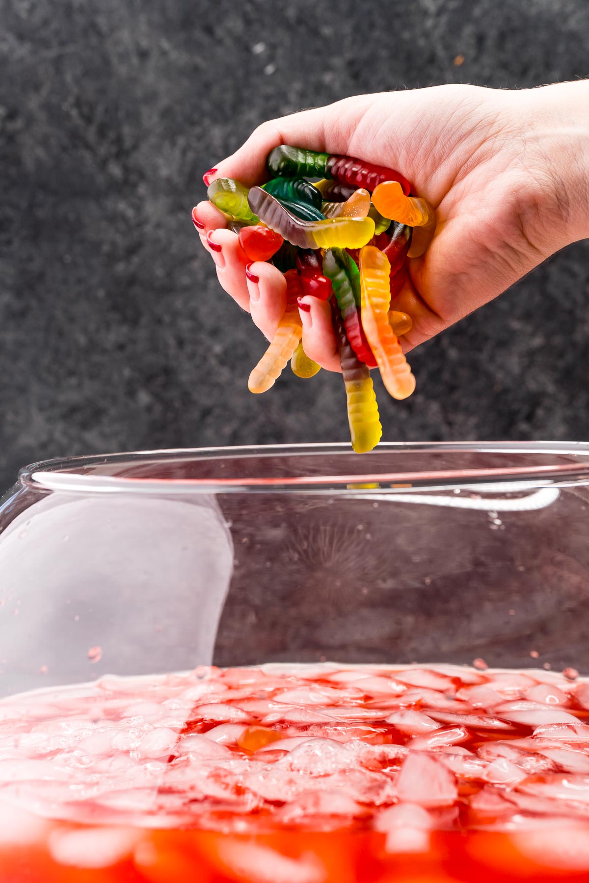 A woman's hand filled with gummy worms over a punch bowl filled with red punch ready to drop them in.