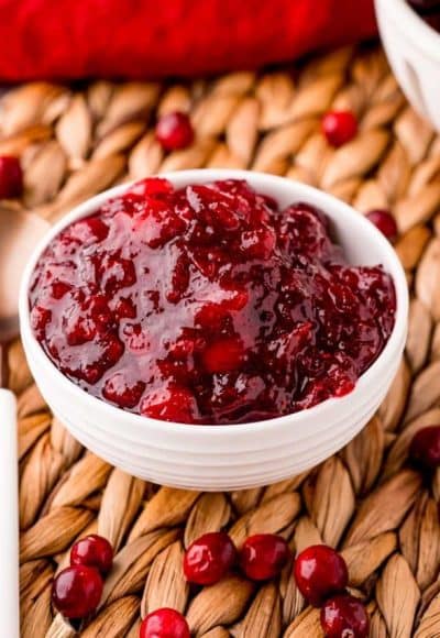 Close up photo of a what bowl filled with cranberry sauce on a whicker placemat with cranberries scattered around it.