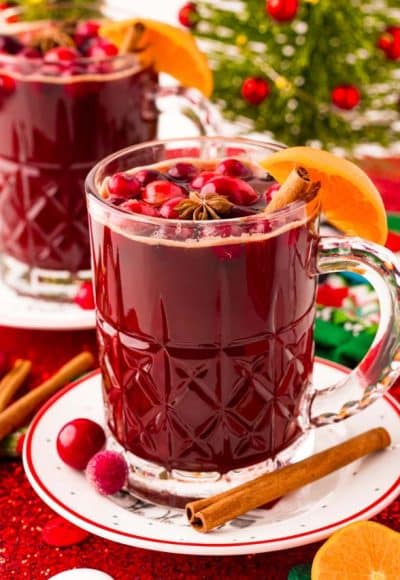 Two mugs of Kinder Punch on saucers with cranberries, oranges, and cinnamon sticks scattered around. Holiday decor in the background.