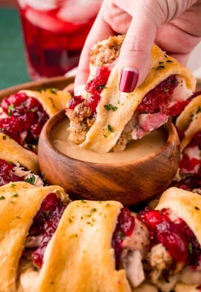 Close up photo of a woman's hand dipping a piece of Thanksgiving Crescent Ring in a small wooden bowl of gravy.