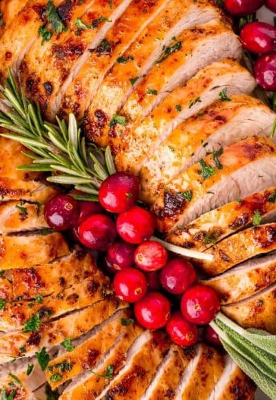 Overhead photo of two sliced turkey tenderloins on a plate with fresh herbs and cranberries for garnish.