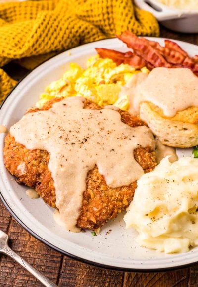 Country friend steak on a white plate with mashed potatoes, eggs, bacon, and a biscuit.