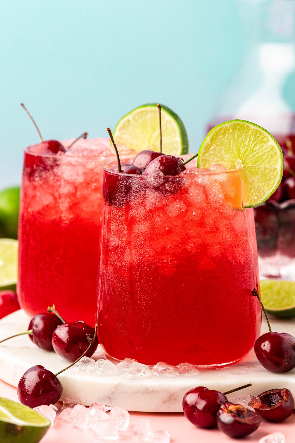 Two glasses of cherry limeade on a table with ice, cherries, and limes around.
