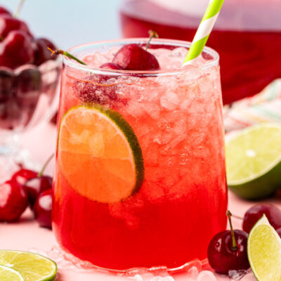 A glass of cherry limeade on a table surrounded by cherries and limes.