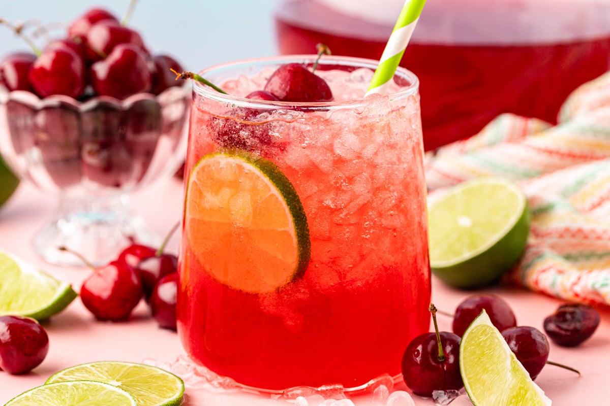 A glass of cherry limeade on a table surrounded by cherries and limes.
