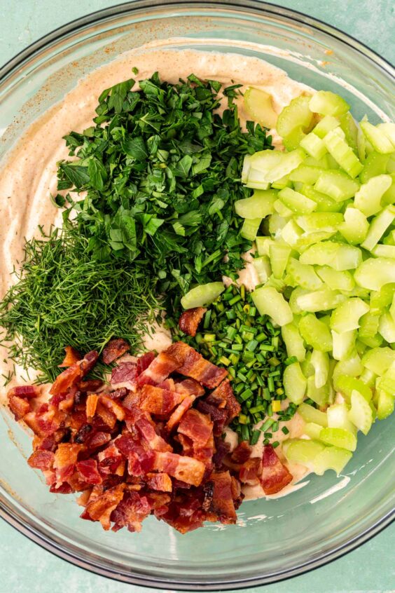 herbs, bacon, and celery in a bowl with dressing for potato salad.