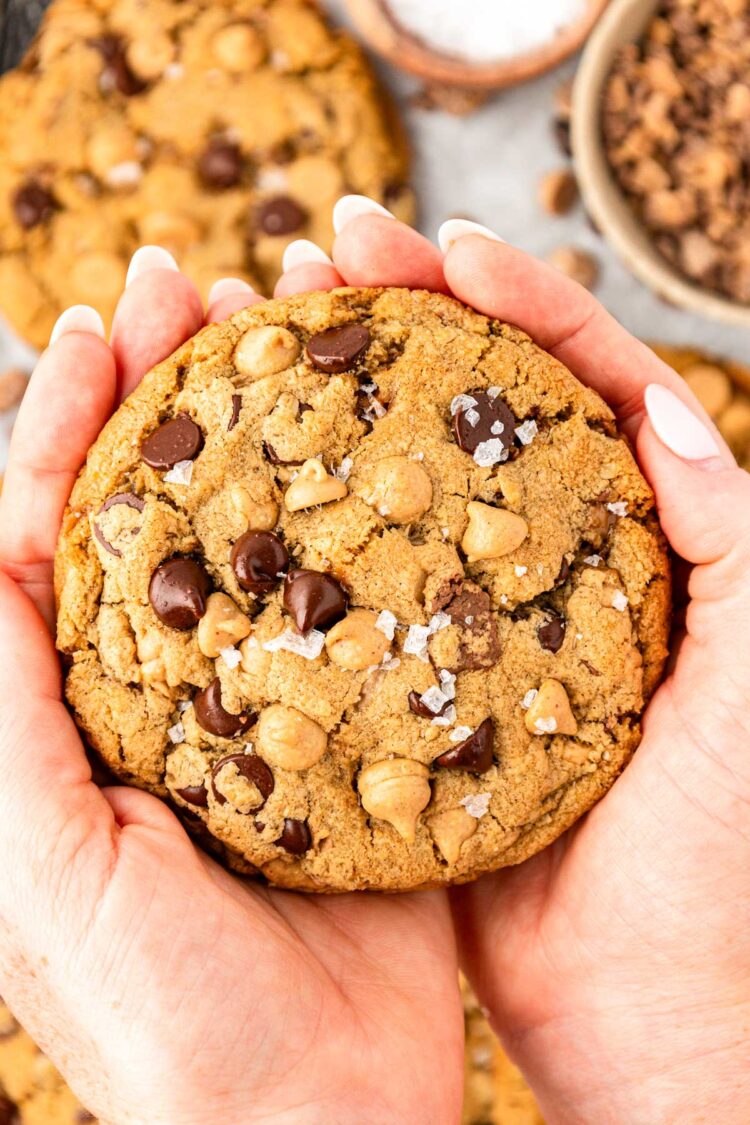 A woman's hand holding a chocolate chip cookie with oatmeal, peanut butter chips, and toffee in it.