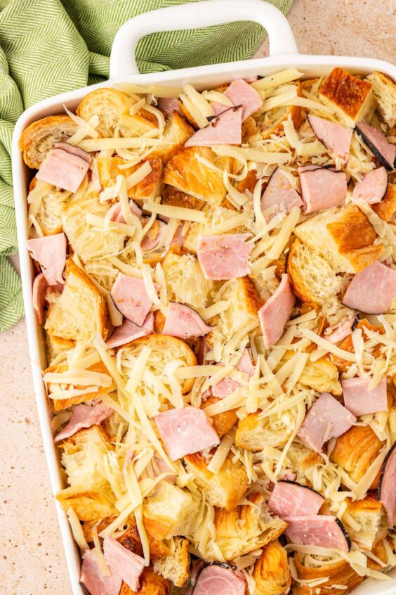 Croissant pieces, ham slices, and shredded cheese in a baking dish.