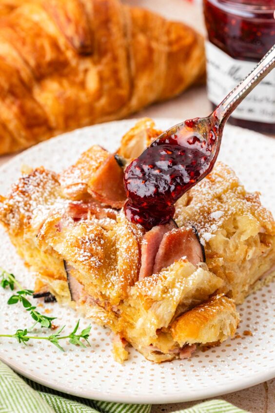 Raspberry jam being spooned over a slice of ham and cheese breakfast casserole on a white plate.