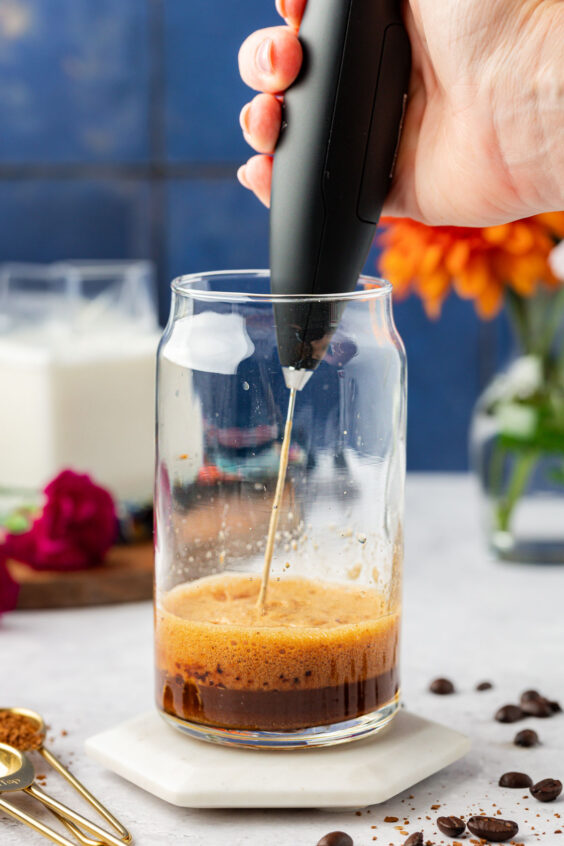 A hand frother whipping water, sugar, and instant coffee in a glass.