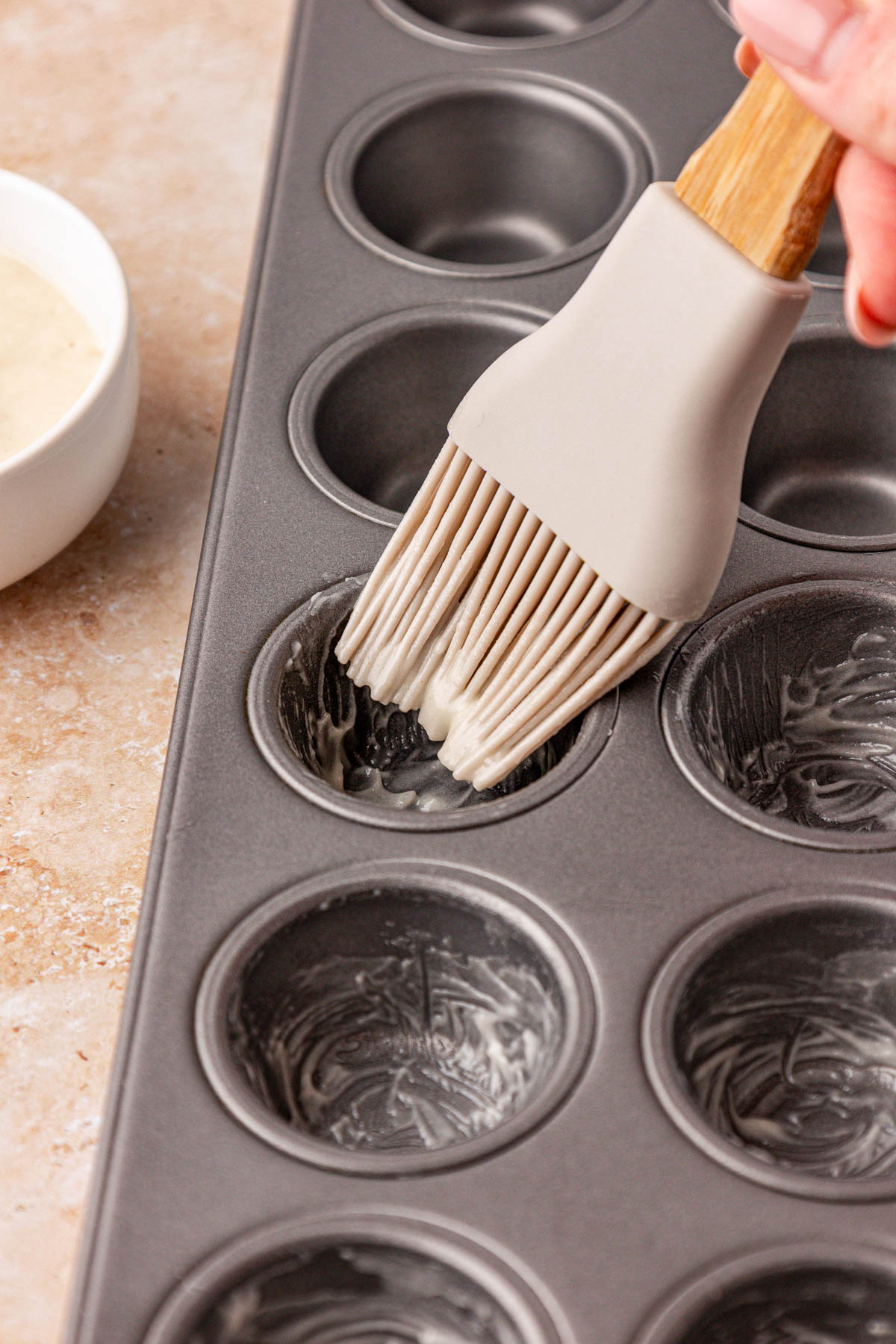 A silicone pastry brush coating the inside of mini muffin tin wells with nonstick paste.