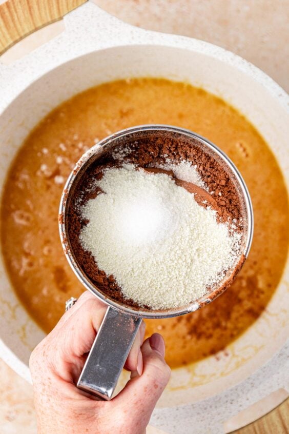 A sifter sifting flour and cocoa into butter and sugar in a pot to make brownie batter.