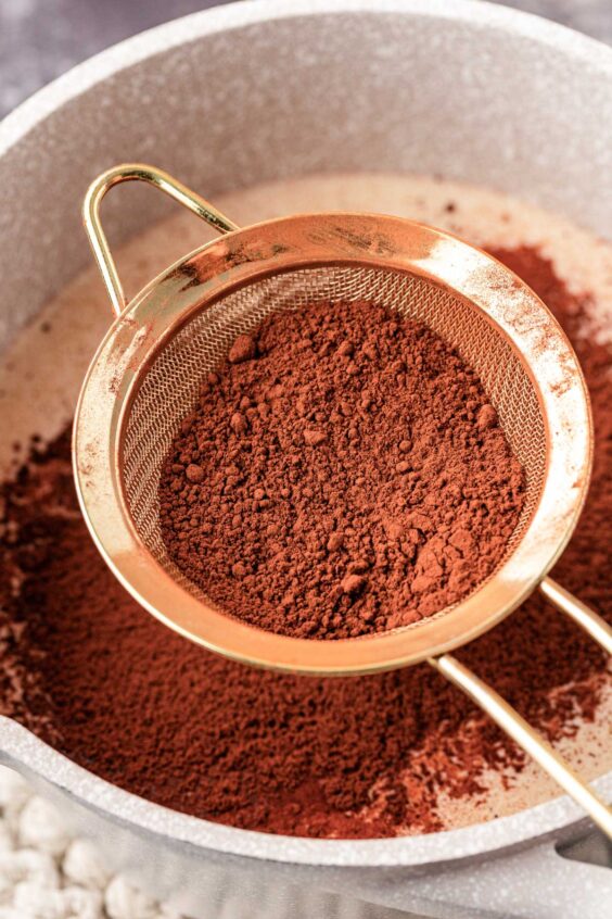 Cocoa powder being sifted into a pot of milk.