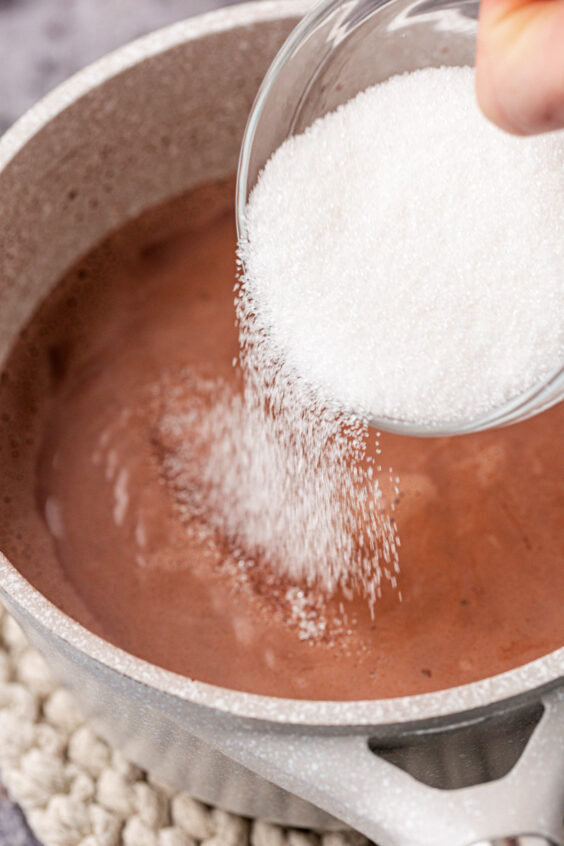 Sugar being poured out of a bowl into a pot of hot chocolate.